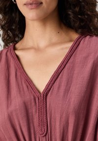 Tunique rose avec un décolleté en V et une finition tressée. Tissu texturé avec un drapé doux et fluide. Gros plan sur les détails du col.