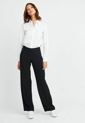 Chemise blanche à manches longues, associée à un pantalon noir large avec six boutons décoratifs noirs à la taille. Baskets blanches.