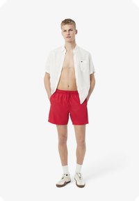 Un short de bain rouge avec une taille élastique, une chemise en lin blanche à boutons, et des chaussettes blanches avec des chaussures présentant un design beige et marron.