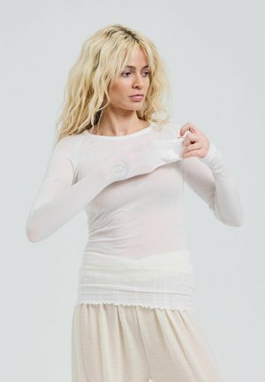 Long-sleeve, sheer white top with a fitted style. Lightweight fabric with a smooth texture. Model adjusts the sleeve, wearing cream pants.