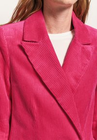 Blazer in corduroy rosa con ampi revers e texture a coste verticali. Presenta un design a un solo petto e una camicia bianca visibile sotto.