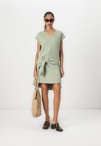 Abito corto verde chiaro con vita annodata, orlo a campana e scollatura a V. Indossato con occhiali da sole scuri, una borsa intrecciata e scarpe con tacco dalla texture.