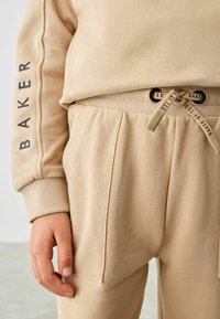 Beige sweatshirt med teksturerede ærmer og "BAKER" trykt langs armen, parret med matchende bukser med snor og plisserede lommer.