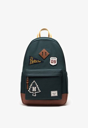 Sac à dos vert foncé avec des accents bruns, une poignée jaune et des écussons brodés dont « Herschel », le numéro 09 et un arbre avec un « H. »