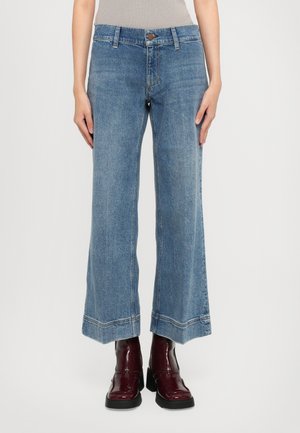 Femme portant un jean large et court bleu et des bottes cheville rouges foncées et brillantes, se tenant devant un fond blanc uni.