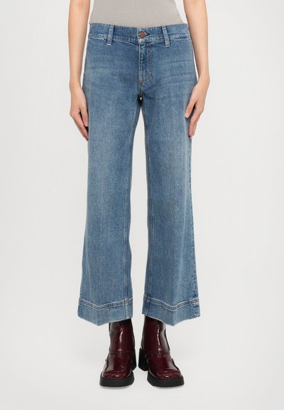 Femme portant un jean large et court bleu et des bottes cheville rouges foncées et brillantes, se tenant devant un fond blanc uni.