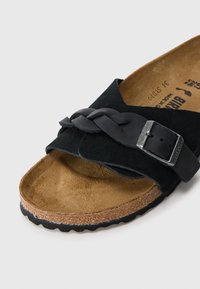Sandale en suede noir avec une sangle tressée et une boucle métallique. Semelle en liège avec une surface texturée marron et une semelle en caoutchouc dotée d'un motif antidérapant.