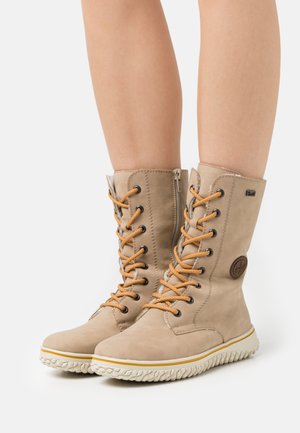 Beżowe buty za kostkę z pomarańczowymi sznurówkami, cholewką z teksturowanej skóry i białą gumową podeszwą. Zawierają naszywkę z logo po boku.