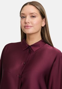 Burgundy satin blouse with a classic collar, featuring button closure and smooth texture. Tailored fit with a slightly relaxed silhouette.