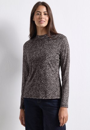 Kvinde med langt brunt hår iført en tætsiddende grå leopardpræget langærmet top og mørkeblå jeans, der står mod en ensfarvet hvid væg.