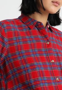 Röd rutig skjorta med blå och vit detalj, med knappstängning fram, krage och korta ärmar. Texturerat tyg med rutig mönstring.