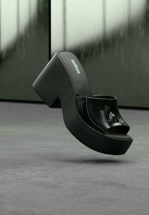 Μαύρο πλατφόρμα κοντό πέδιλο με γυαλιστερό λουράκι ανοιχτής μύτης και το λογότυπο "Melissa" στην εσωτερική σόλα, τοποθετημένο πάνω σε θολό εσωτερικό φόντο.