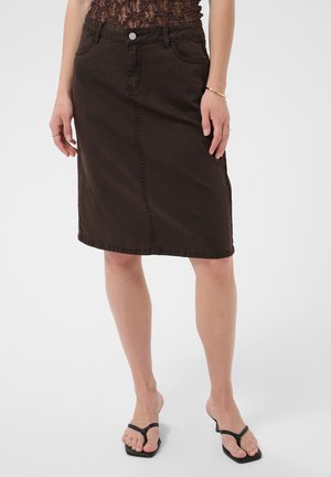 Personne portant une jupe en jean marron foncé jusqu'aux genoux, des tongs noires, un haut en dentelle et des bijoux simples, debout devant un fond uni.