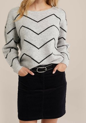 Vrouw die een lichtgrijze trui draagt met zwarte zigzaglijnen en een zwarte corduroy rok met een zwarte riem met zilveren gesp.