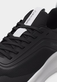 Μαύρο αθλητικό παπούτσι με μαύρα κορδόνια, λευκή υφή στη σόλα και λευκή γλώσσα με το λογότυπο Calvin Klein.