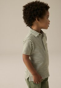 Grøn og hvidstribet polo-shirt med krave og korte ærmer, parret med grønne bukser. Modellen står i profil mod en neutral baggrund.
