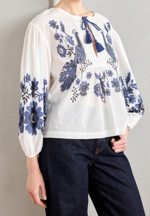 Witte blouse met blauwe bloem- en pauwembroidery, lange pofmouwen, halslijn met kwastjes en strik, gedragen met donkere spijkerbroek.