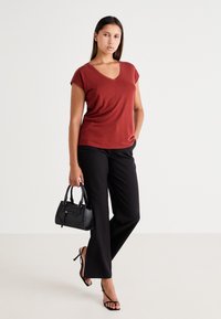 Roestkleurig T-shirt met korte mouwen en een V-hals, gecombineerd met zwarte wijde broek. Zwarte handtas met korte hengsels en slanke sandalen. Soepele texturen.