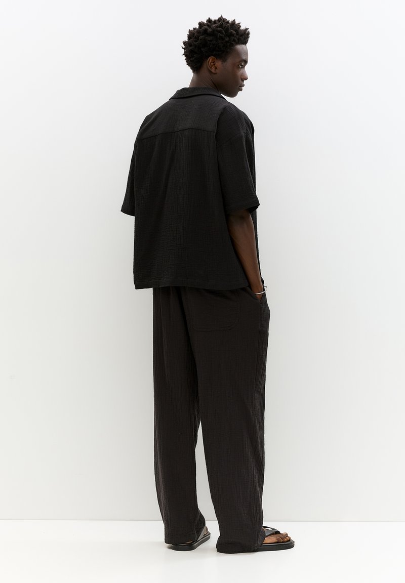 Camicetta nera a maniche corte con texture e vestibilità comoda, abbinata a pantaloni neri larghi. Presenta un colletto e tasche laterali. Infradito completano il look.
