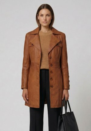 Kvinde iført en brun læderjakke med knapper over en strikket sweater og sorte bukser, der holder en stor sort lædertaske.