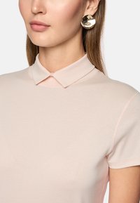 Light pink polo shirt with a structured collar, smooth texture, and contrasting stitching. Accompanied by large metallic round earrings.