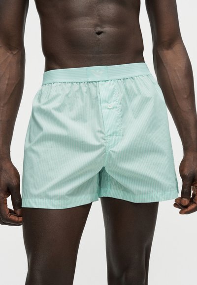 Ανδρικό μοντέλο φορώντας φωτεινά τυρκουάζ ριγωτά μποξεράκια με κουμπιά και ελαστική μέση σε απλό λευκό φόντο.