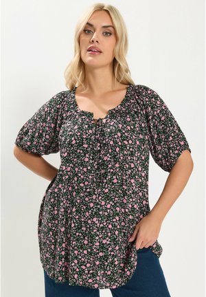 Donna con capelli biondi che indossa una camicetta nera con motivo floreale rosa e maniche corte a sbuffo, in piedi con una mano sul fianco.