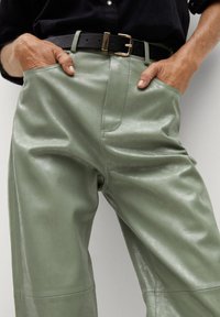 Los pantalones de cuero verdes presentan una cintura alta con un cinturón negro y una hebilla dorada, exhibiendo una textura suave y brillante, además de bolsillos laterales.