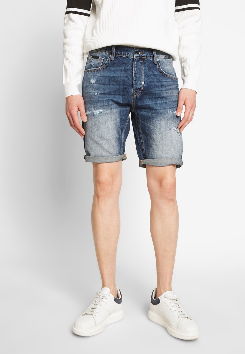 Jeansshorts i urtvättad blått med slitna detaljer. Upprullad fåll och femficksdesign. Kombinerat med en vit ribbad tröja och vita sneakers.