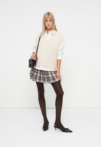 Młoda kobieta w beżowym bezrękawniku na białej koszuli, w spódnicy w kratkę, czarnych rajstopach i czarnych butach na obcasie typu slingback, nosząca czarną torebkę na ramię.