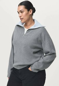 Mujer con el cabello oscuro recogido en un moño, vistiendo un suéter gris con cremallera sobre una camisa blanca, con una mano en el bolsillo de unos pantalones negros, mirando hacia la izquierda.