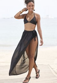Čierny bikini s trojuholníkovým vrškom a vysokým priesvitným sukňou s bočným rozparkom. Doplnkové sandále a vrstvené zlaté náhrdelníky.