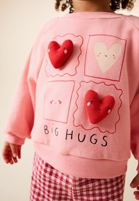 Felpa rosa con due applicazioni a forma di cuore rosso e scritta "BIG HUGS". Tessuto morbido, polsini a costine e vestibilità comoda. Abbinata a pantaloni a quadri.