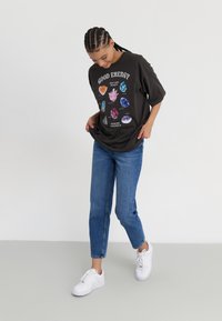 T-shirt surdimensionné gris foncé avec le texte "GOOD ENERGY" et des illustrations de cristaux, associé à un jean bleu et des baskets blanches.