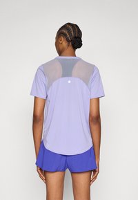 Μωβ αθλητική μπλούζα με πλέγμα στην πλάτη. Κοντά μανίκια, στρογγυλεμένο τελείωμα και ανακλαστική λεπτομέρεια στην πλάτη. Συνδυασμένη με μπλε σορτς.