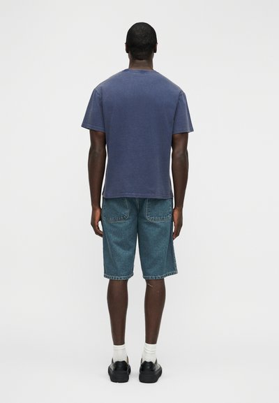 Σκούρο μπλε βαμβακερό μπλουζάκι συνδυασμένο με ανοιχτόχρωμα τζιν σορτς. Καθημερινά παπούτσια με άσπρες κάλτσες, σε προβολή από πίσω με φόντο μια απλή επιφάνεια.