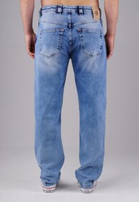 Mann, der hellblaue verwaschene Jeans und weiße Converse-Sneaker trägt, steht mit dem Rücken zur Kamera vor einem einfarbigen Hintergrund.
