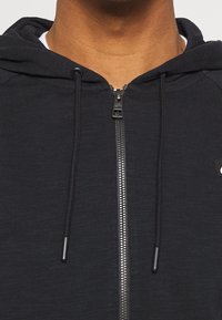 Svart zip-up hoodie med texturerat tyg, dragsnören och framzipper, med en liten logga på bröstet och ribbade ärmslut.