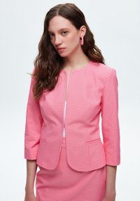Blazer a quadri rosa con scollo tondo, maniche a tre quarti e chiusura con zip frontale. Il tessuto è texturizzato e sagomato.