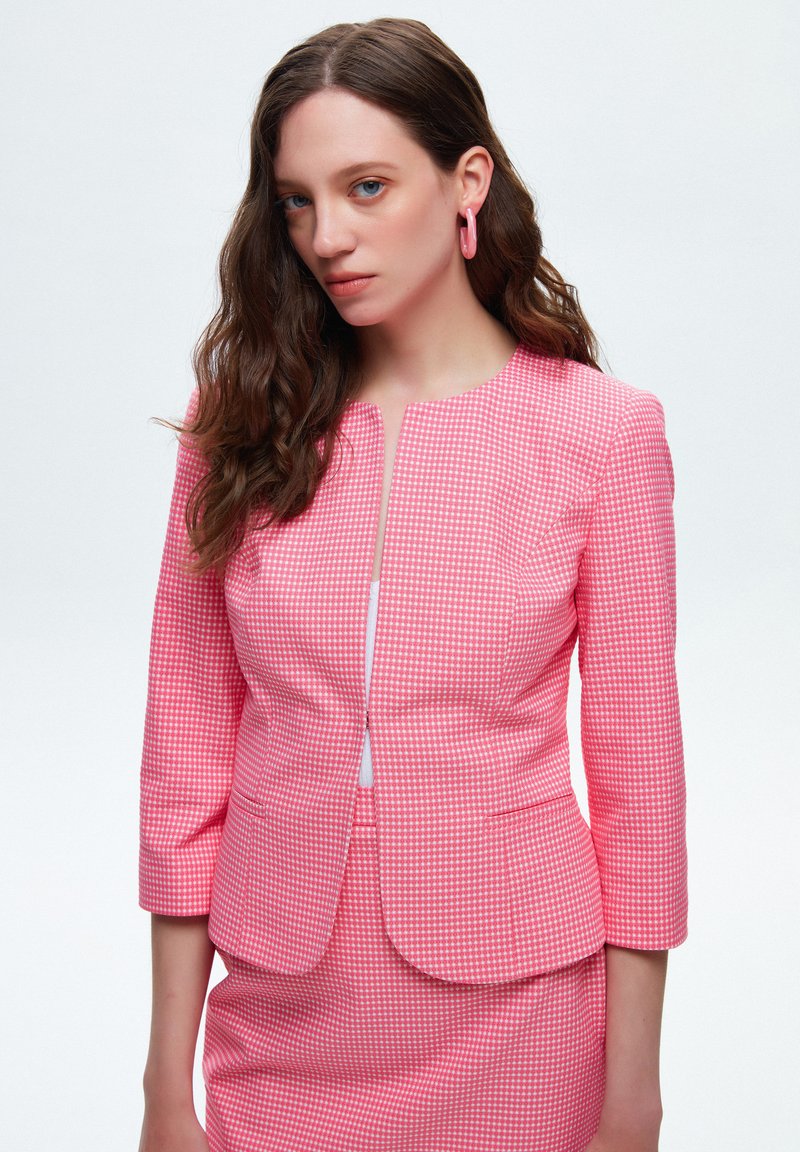 Blazer a quadri rosa con scollo tondo, maniche a tre quarti e chiusura con zip frontale. Il tessuto è texturizzato e sagomato.