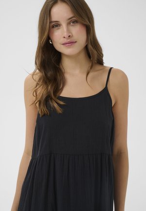 Joven con cabello largo y castaño, vestida con un vestido negro sin mangas con tirantes delgados, sobre un fondo claro y liso.