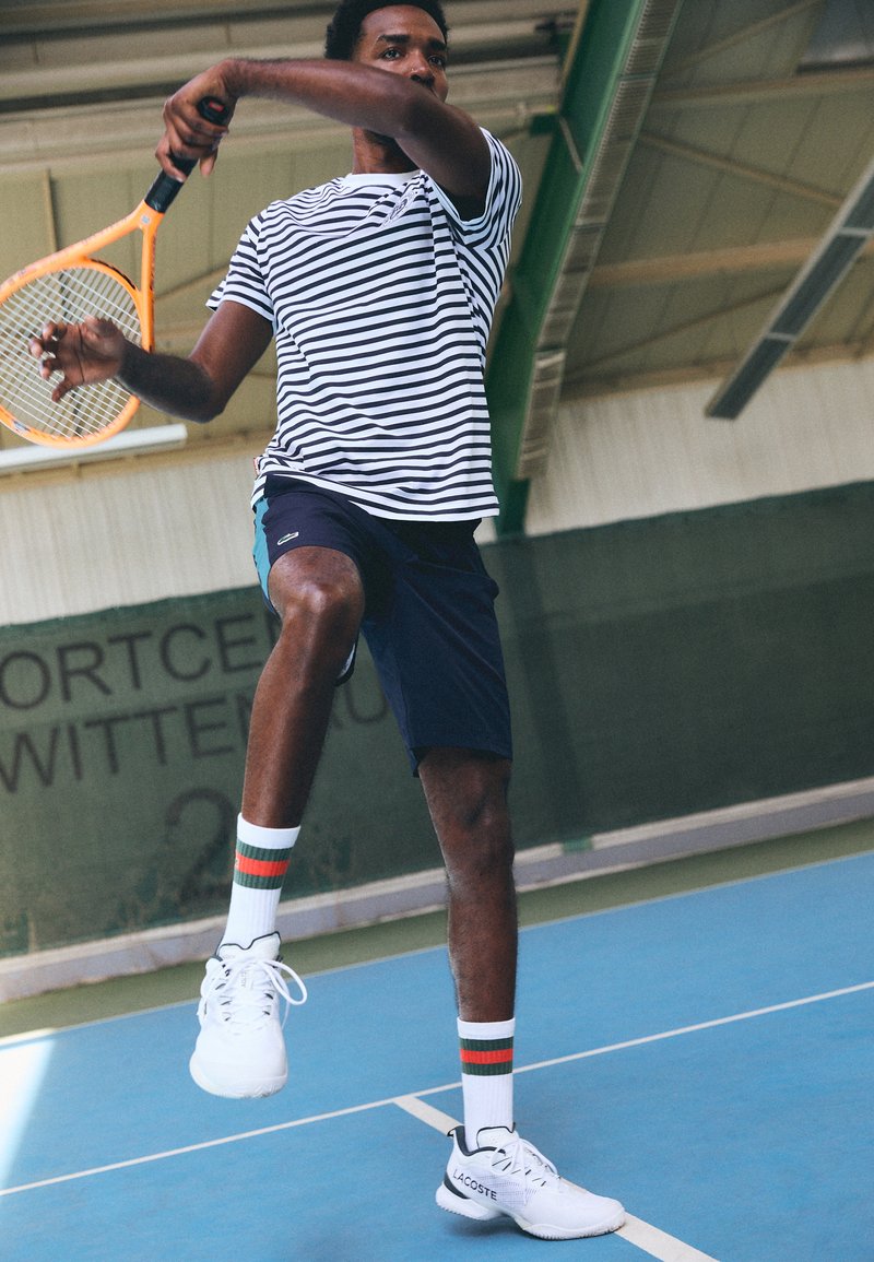 Joueur de tennis sportif portant une chemise à rayures à manches courtes, un short marine, des baskets blanches et des chaussettes à motifs, prêt à frapper une balle de tennis à l'intérieur.