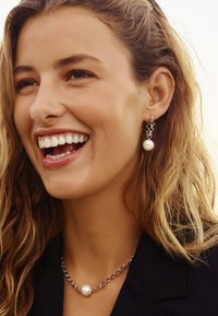 Silverkedjehalsband med ett hängande pärlberlock; matchande pärlörhängen. Motiv har vågigt hår och ett brett leende.