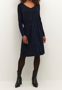 Tmavě modré, dlouhé šaty s výstřihem do V, elastickým pasem a mírně rozšířenou sukní. Nosené s černými kotníkovými botami.