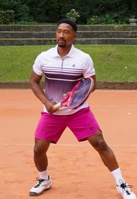 Un joueur de tennis masculin portant un polo blanc et gris avec un short rose, tenant une raquette de tennis, sur un court en terre battue avec des alentours verdoyants.