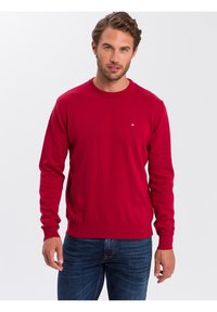 Czerwony sweter z dzianiny z okrągłym dekoltem, prążkowanymi mankietami oraz wykończeniem. Posiada małe logo na klatce piersiowej. Połączony z ciemnoniebieskimi dżinsami.