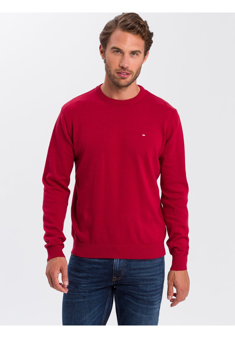 Czerwony sweter z dzianiny z okrągłym dekoltem, prążkowanymi mankietami oraz wykończeniem. Posiada małe logo na klatce piersiowej. Połączony z ciemnoniebieskimi dżinsami.
