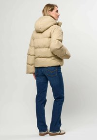 Femme de profil portant une doudoune beige à capuche, un jean bleu et des baskets blanches sur un fond clair uni.