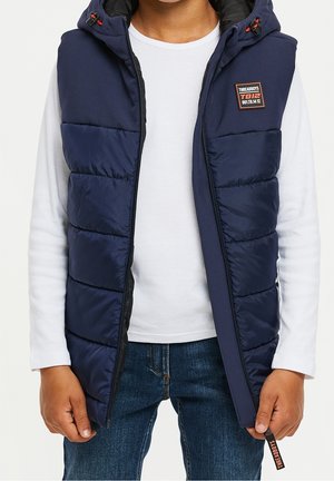 Granatowa pikowana kamizelka z kapturem, z przeszywanymi panelami, zapięciem na zamek i naszywką z logo. Noszona na białej koszuli z długim rękawem.