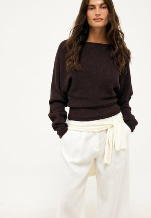 Kvinde med langt, bølget hår iført en mørkebrun langærmet sweater og hvide bukser med højtalje og stofrem bundet i taljen.